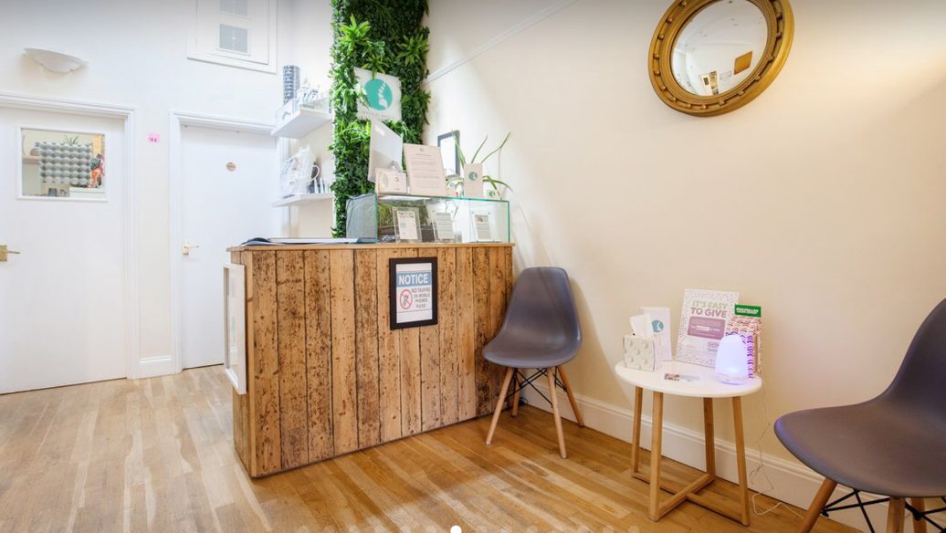 The clinic reception: a reclaimed timber desk in front of a living plant wall, with waiting chairs and a small side table.