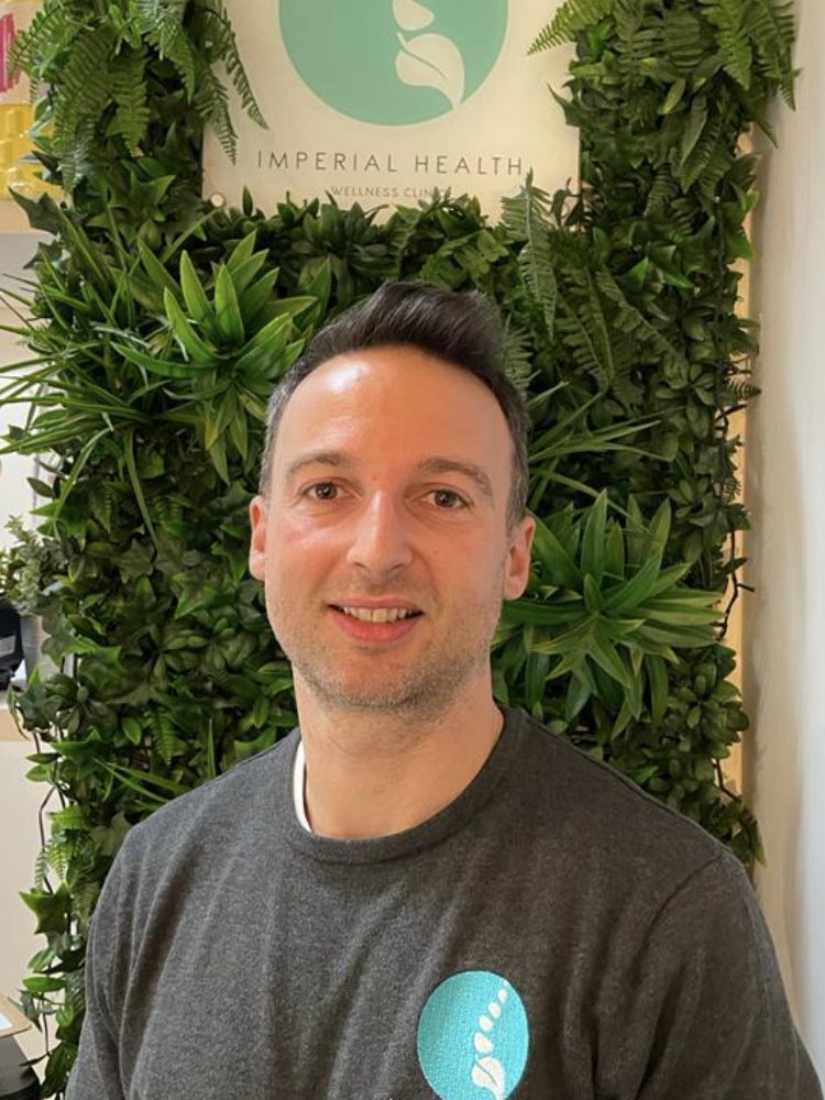 Toby Wragg in a grey Imperial Health top, standing in front of the clinic's living plant wall.